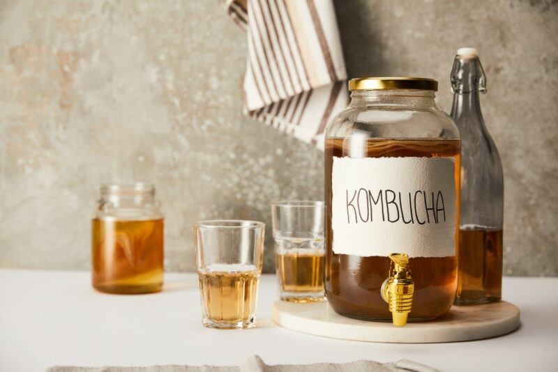 A large bottle of kombucha brewing next to a smaller bottle and a few glasses of kombucha tea