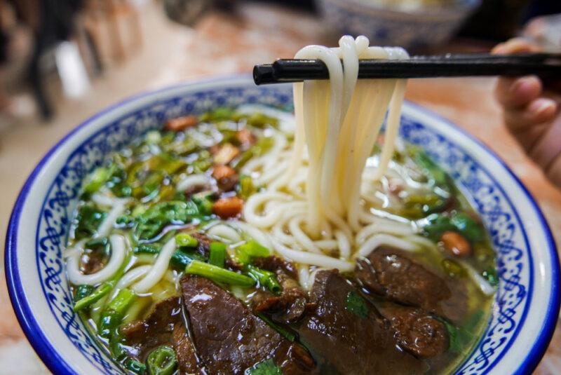 a closeup image of a white bowl with blue prints containing lamian noodles with wooden chopsticks