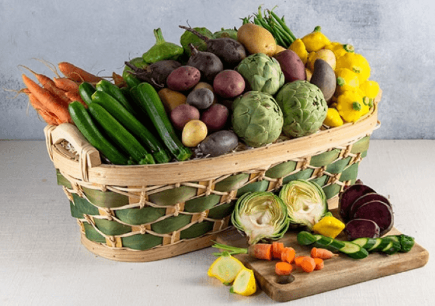 Baby vegetable basket.  The basket is a tan and green weaved basket filled with baby vegetables such as carrots, zucchini, tricolor potatoes, artichokes.  IN front of the basket is a wooden cutting board with sliced vegetables