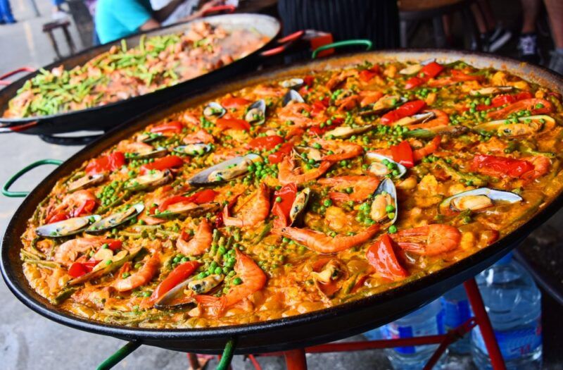 Two large dishes of paella being sold at a street market. They each have many toppings, including various types of seafood