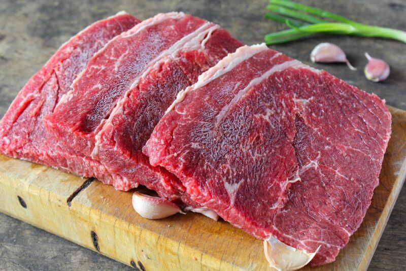 A wooden board with large thick slices of top round steak next to some garlic cloves