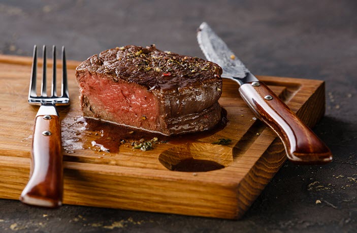 A cutting board with a rare steak, a knife and fork