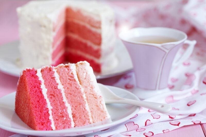 a white ceramic dish with a slice of layer cake in different shades of pink, at the back is the rest of the layer cake with a cup of tea beside it