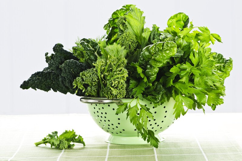 This photo shows several types of green leafy vegetables in a silver colander on a green and white cloth.