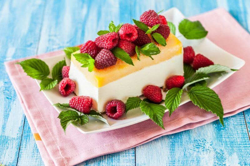 A layered lemon semifreddo dessert with a white base, yellow top, that is surrounded by raspberries and raspberry leaves