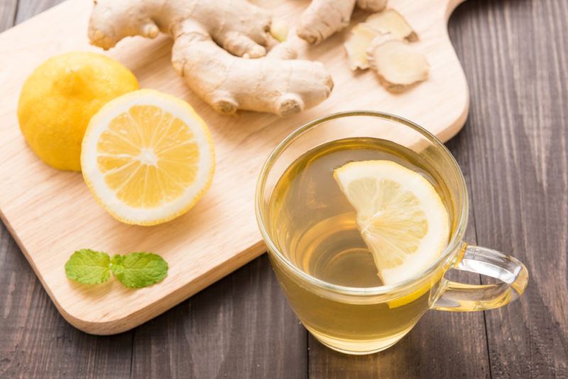 A glass mug of lemon ginger tea with a lemon slice, next to a cutting board containing lemon and ginger.