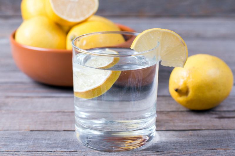 A glass of lemon water with a bowl of lemons and individual lemons in the background