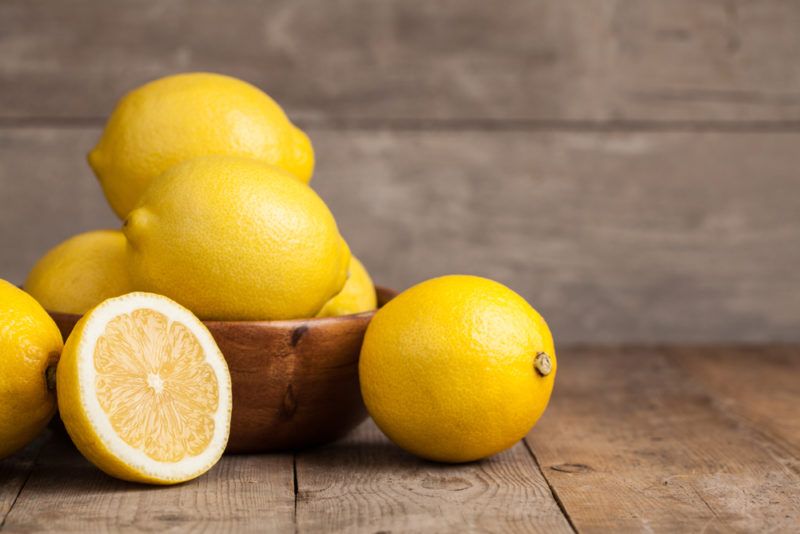 A small wooden bowl with whole lemons, along with a whole lemon and a lemon half on a table