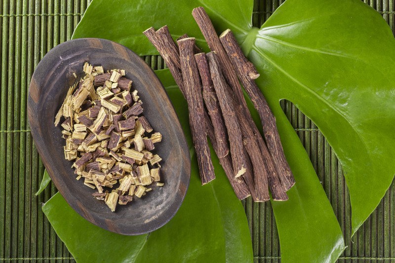 This photo shows an overhead view of several pieces of chopped licorice in a wooden bowl and several pieces of whole licorice root beside it on the right, on a large green leaf, which is sitting on a green mat.