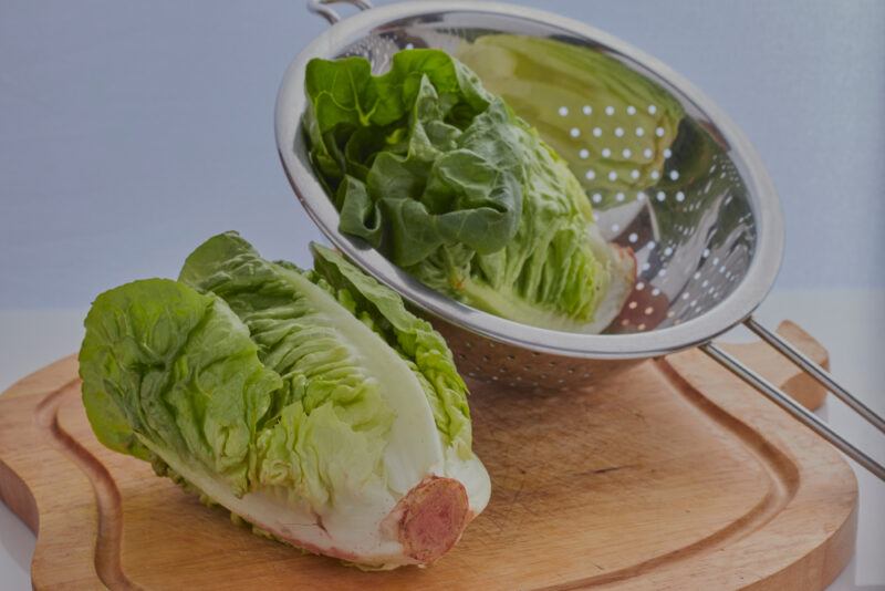 on a wooden chopping board is a head of Little Gem lettuce and a metal colander with Little Gem lettuce as well