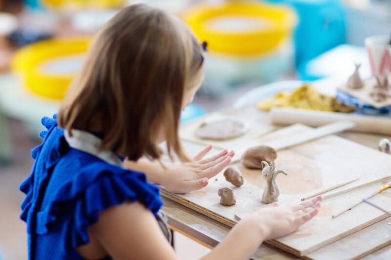 Craft of the Month Club for Kids - young girl at table working with clay shaping small figurines such as a cat