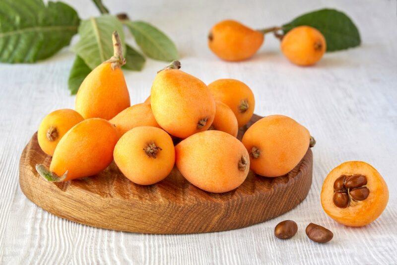 A wooden board containing a pile of loquat fruits. There are a few more scattered on the white table