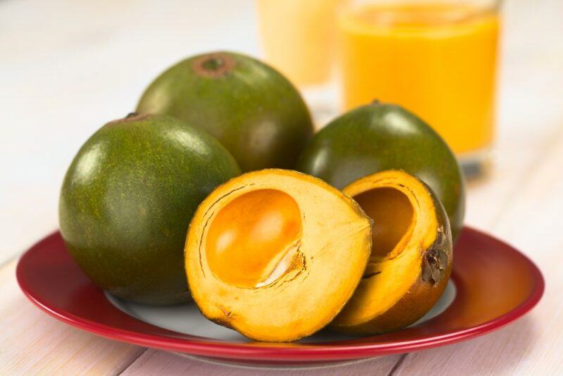 Three full lucuma fruits on a red plate with one that's been cut in half and a few glasses in the background