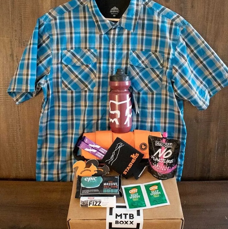 MTB Boxx blue plaid shirt in the background with an MTB Boxx in front with cycling products on top such as water bottle, deep woods off, can koozie, and more