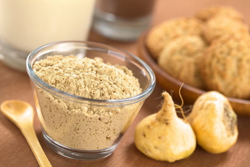 A glass bowl of maca powder next to a spoon and maca roots