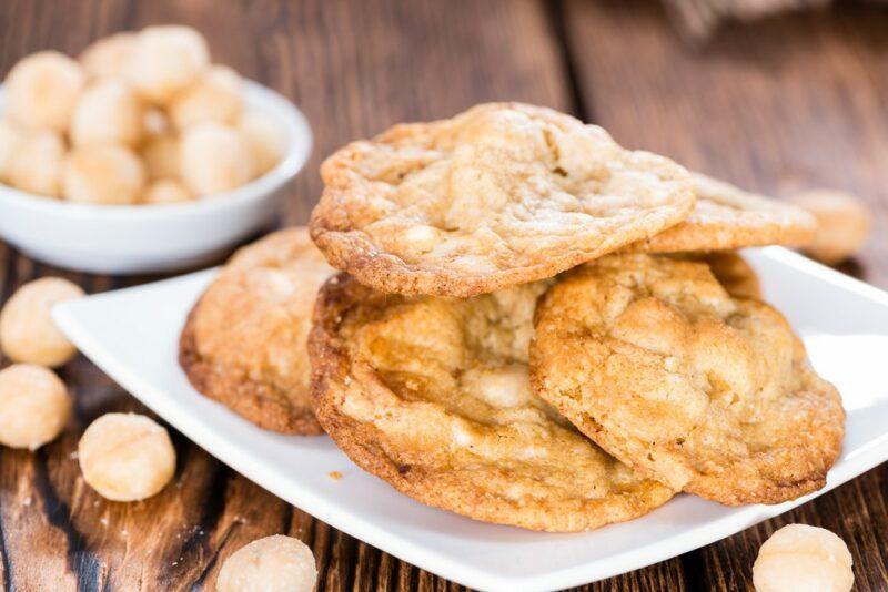 A white plate with a stack of macadamia nut cookies, with some nuts on the table, and another small bowl of them behind the plate