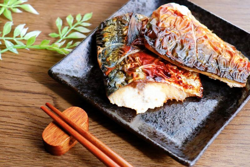 Saba,No,Sio,Yaki,(grilled,Mackerel).,A,Traditional,Japanese,Grilled | Food For Net This photo shows two pieces of grilled mackerel on a dark square plate on a wooden table next to chopsticks and green leaves.
