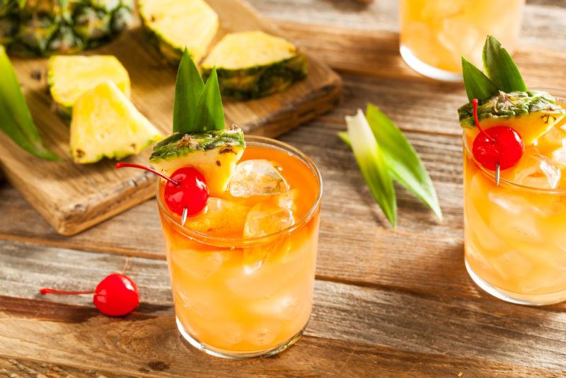 Three mai tai cocktails on a table with cherries, pineapple and leaves