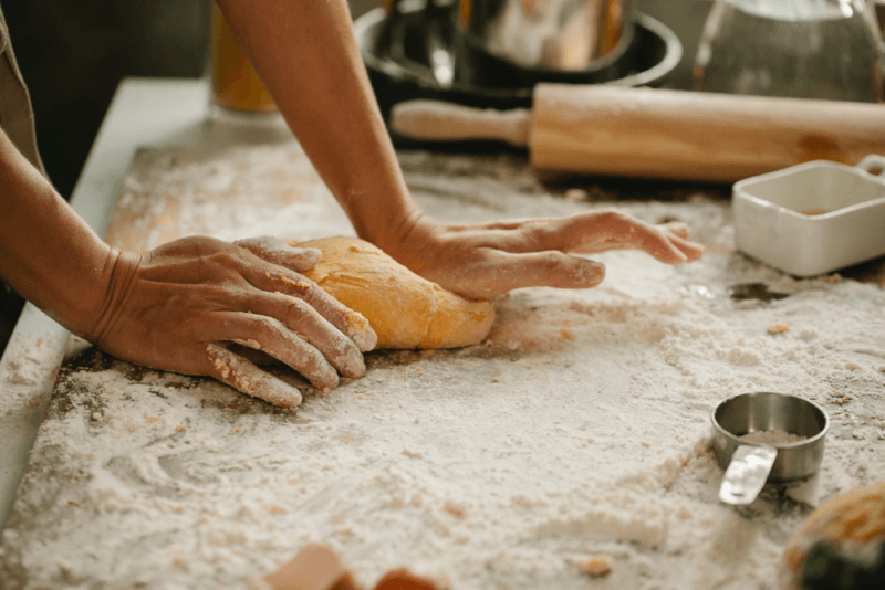 Someone rolling out dough on a table. There is bread flour scattered on the table and the dough has been made with this same flour.