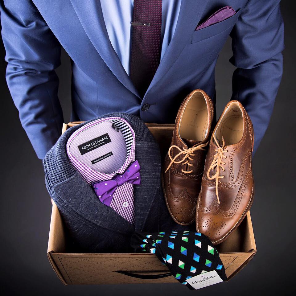 Man in suit holding an Urbane Box inside the box is a cardigan, with purple plaid shop and purple bow tie, brown shoes, and fashion socks