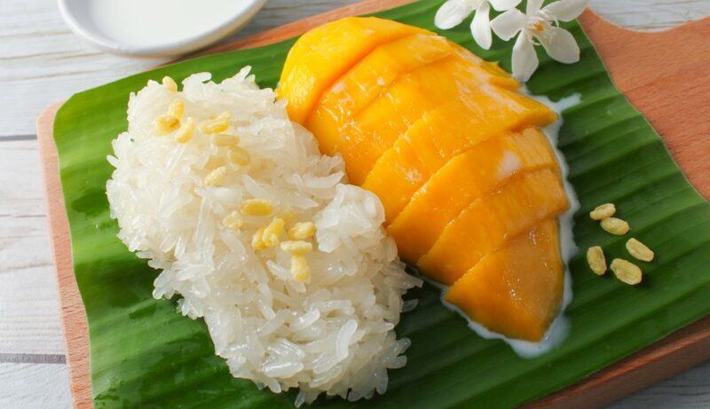 A green leaf with mango sticky rice and sliced mango, next to some flowers