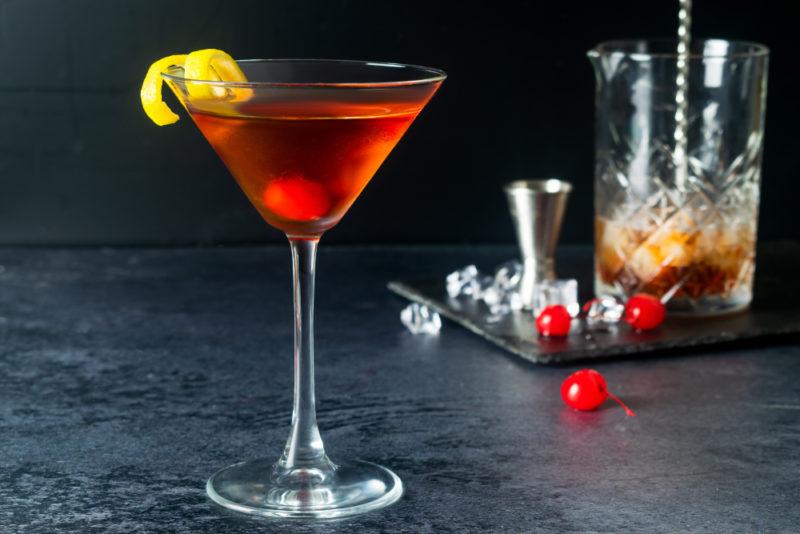 A Manhatten cocktail in a cocktail glass with the various ingredients and implements in the background