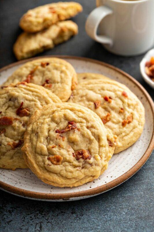 A white and brown plate with four maple bacon pancake cookies, with two cookie halves and a mug in the background