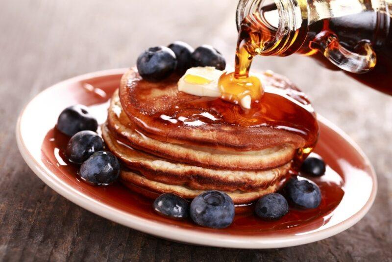 A plate of pancakes with blueberries and butter, where someone is pouring maple syrup on top