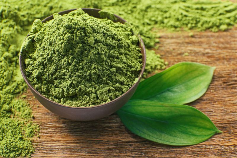 A brown bowl that contains matcha powder, with more matcha powder and a couple of leaves on the table