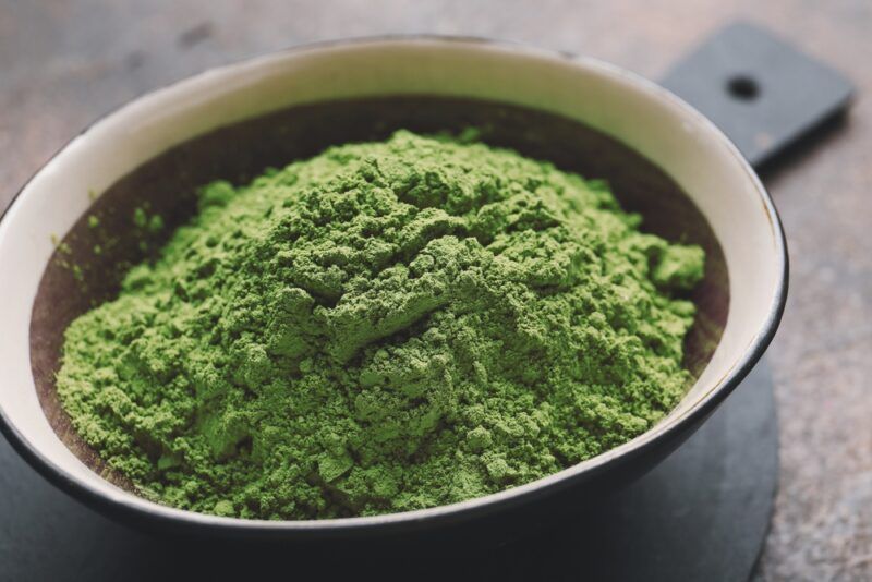 a closeup image of a ceramic brown bowl with a wide cream rim full of matcha, resting on top of a black wooden chopping board