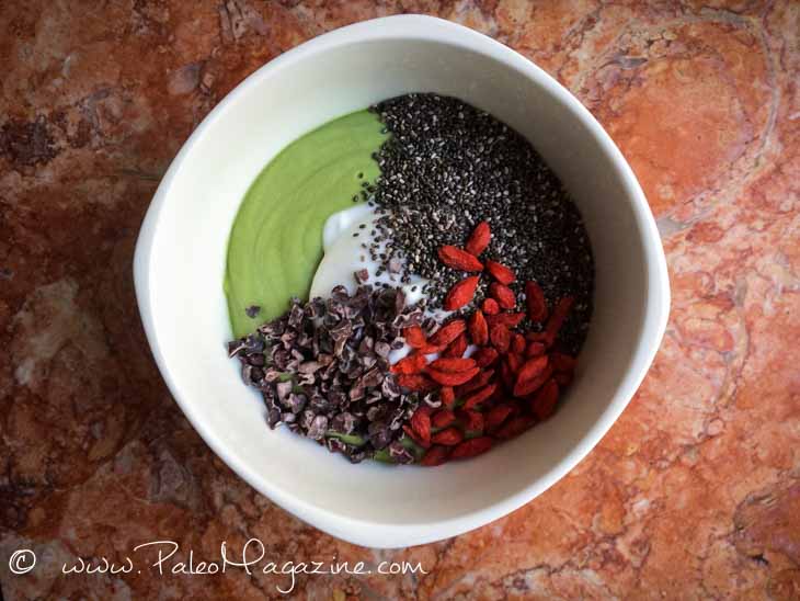 A white bowl with a matcha smoothie, chia seeds, cocoa nibs and goji berries