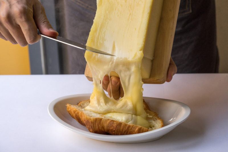 Melted rachlette cheese being scraped onto a croissant