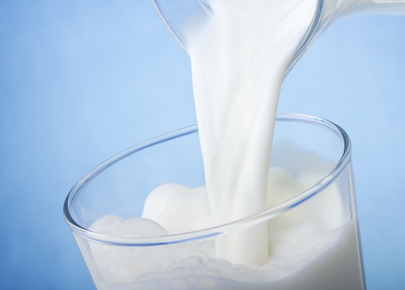 A stream of milk flows from a pitcher into a glass almost full of milk, against a blue background.