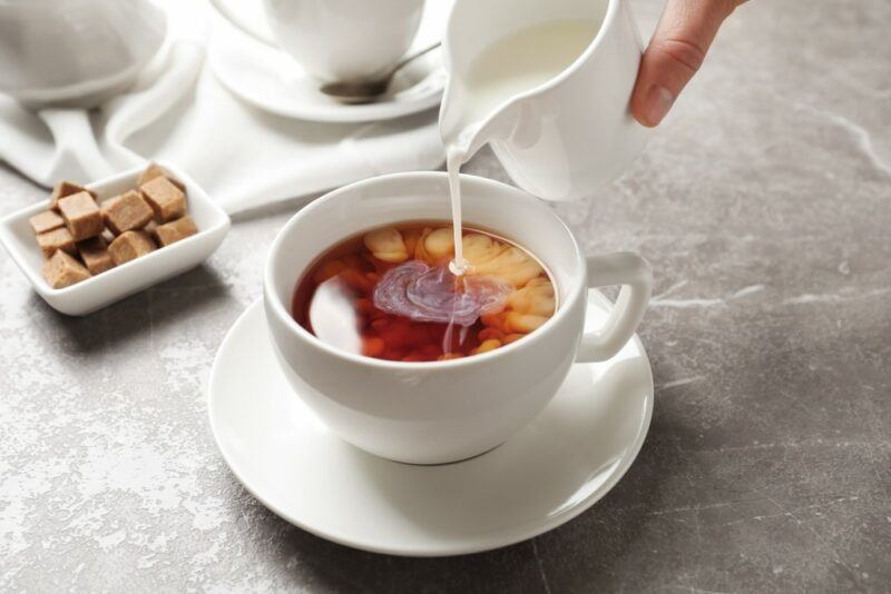 A cup of coffee or tea on a table with milk being poured into it and sugar in the background