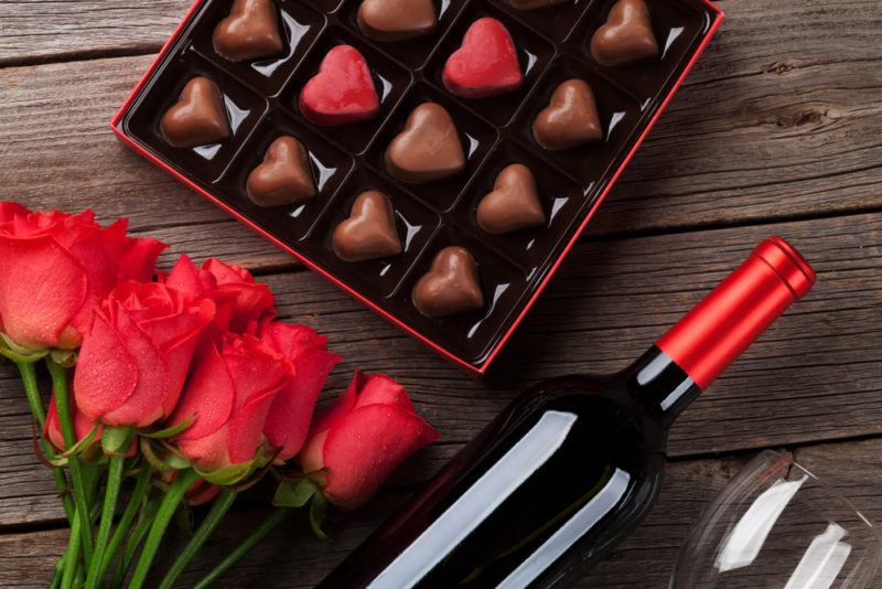 A box of heart shaped milk chocolates, next to a bunch of red roses, a bottle of red wine and a glass