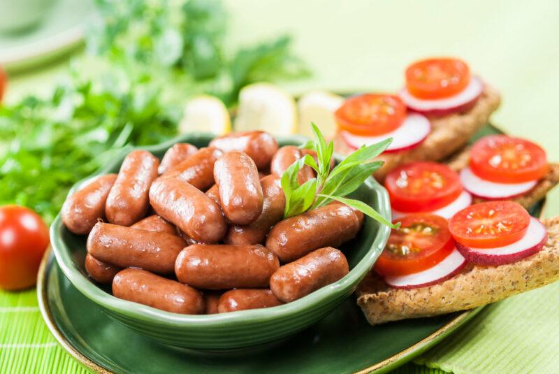 A green dish of brown mini sausages rests on a green plate that also holds bread slices with radishes and tomatoes on them.