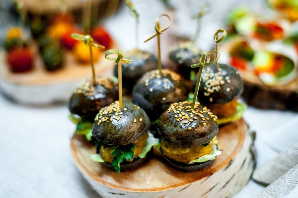 A plate that contains small black burgers, with other appetizers in the background