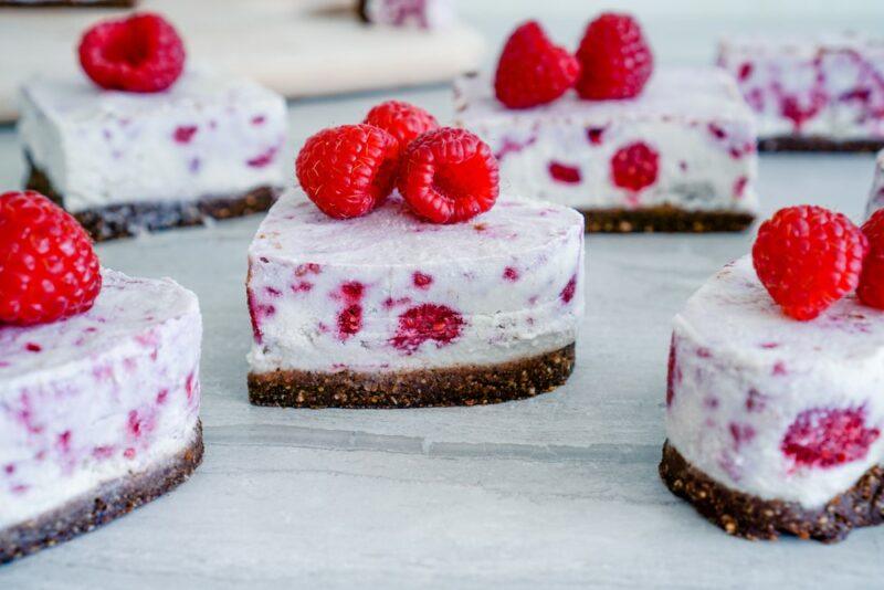 A white table with mini cheesecakes in the shape of hearts, with raspberries in the cheesecake itself and also as a garnish