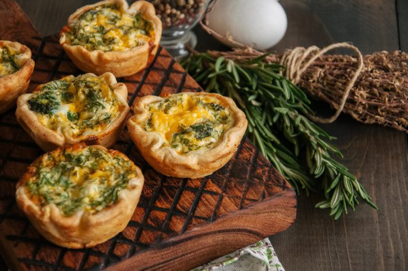 A board that contains five mini quiches next to herbs, an egg and another ingredient