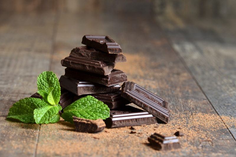 A small pile of dark chocolate next to some mint leaves on a wooden floor with chocolate powder