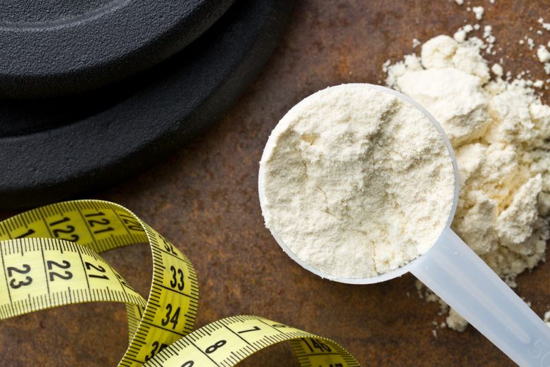 A scoop of white protein powder near a tape measure and a weight