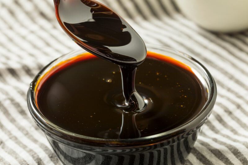 Organic,Black,Cane,Sugar,Molasses,In,A,Bowl | Food For Net A spoon full of molasses drips molasses into a clear glass bowl resting on a gray and white striped cloth.