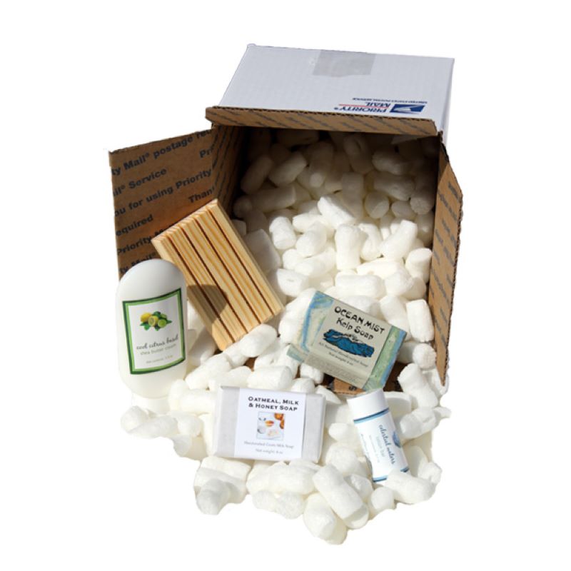 White US postal box with Styrofoam peanuts spilling out and two bars of soap, lotion, and a wooden soap holder, arranged on top.