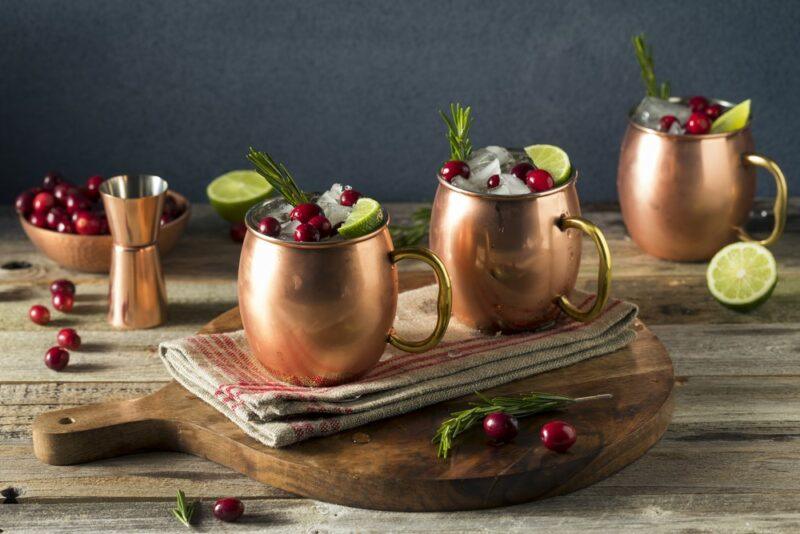 A wooden table with three copper mugs containing an apple cranberry Moscow mule. There are various pieces of lime and cranberries on the table, along with a container of cranberries and a jigger