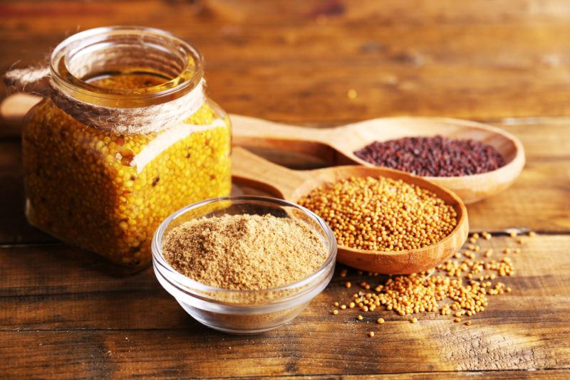 A small glass bowl of mustard powder, a wooden spoon of red mustard seeds, a wooden spoon of yellow mustard seeds and a jar of mustard