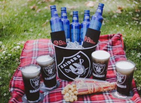 A Raiders bucket on a picnic blanket containing 5 Bud Light bottles. On the blanket are 4 filled Raiders glasses on coasters and a spilled container of popcorn. 