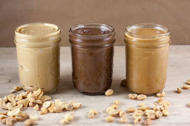 Three jars of nut butters rest on a wooden board with several loose nuts.