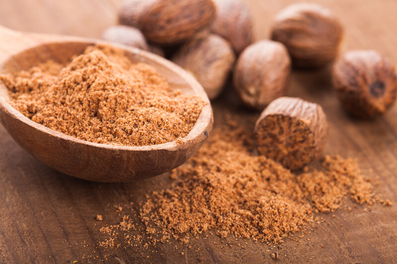 wooden ladle full of ground nutmeg on top of  a wooden surface with whole nutmegs and loose nutmeg powder