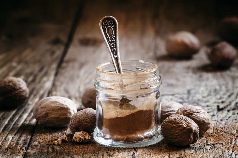 small spice jar with powdered nutmeg and small teaspoon on an aged-wooden table with scattered nutmeg seeds around it