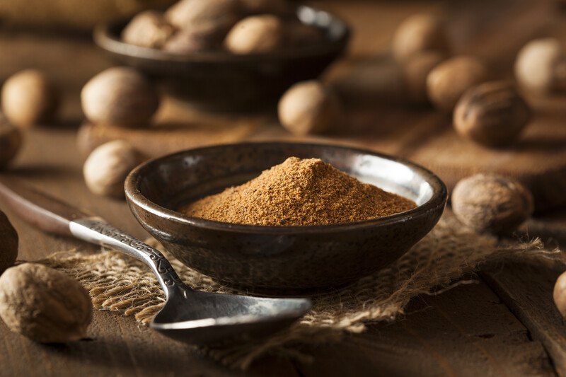 Ground nutmeg in a dark brown clay saucer resting on  a piece of sack cloth, with spoon and scattered nutmeg seeds around it.